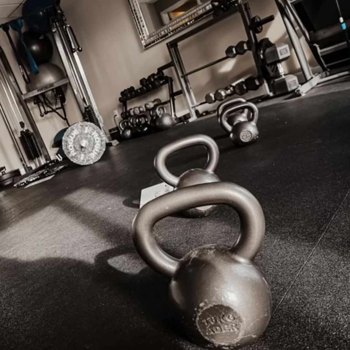 A gym interior with kettlebells on the floor, various exercise equipment, and a mirror reflecting the space.