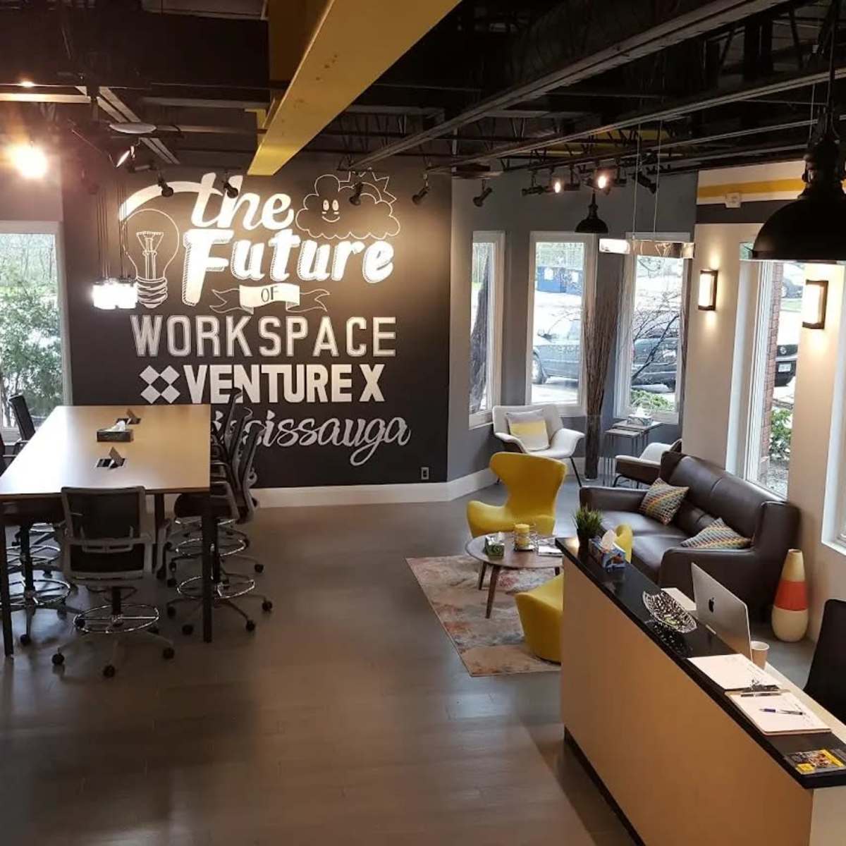 A modern office space featuring a long table, various seating arrangements, and a prominent wall mural that reads "the Future WORKSPACE VENTURE X Mississauga."