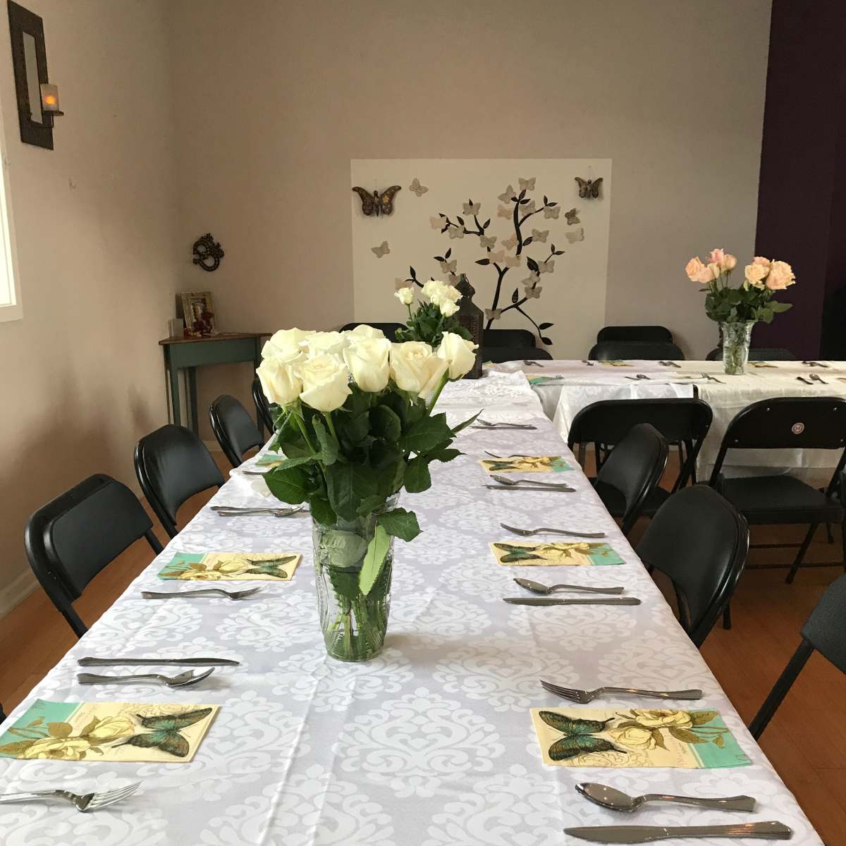 A long, neatly set dining table with white tablecloths, silverware, and a vase of white roses sits in a room decorated with a butterfly-themed wall art.