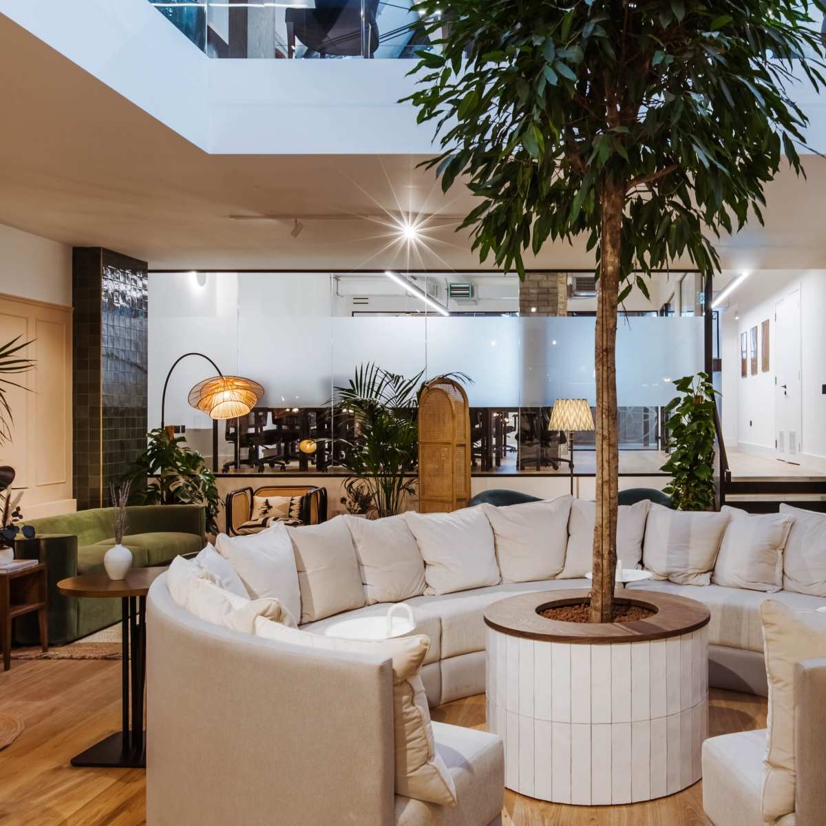 A spacious interior featuring a circular seating area around a central tree, with a kitchen in the background and large windows allowing natural light.