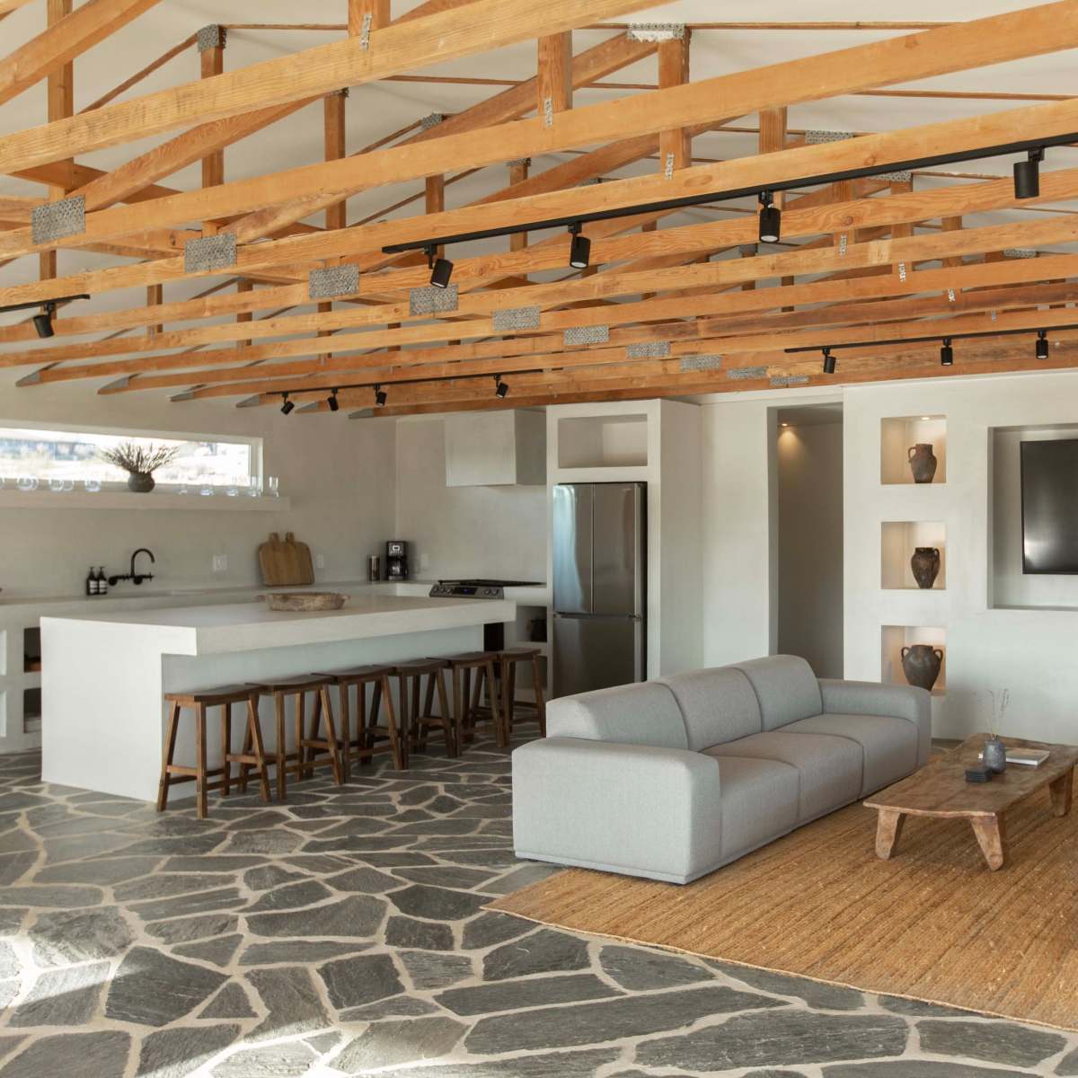 A modern living area featuring a spacious layout with wooden beams, a gray sofa set, stone flooring, and a kitchen in the background.