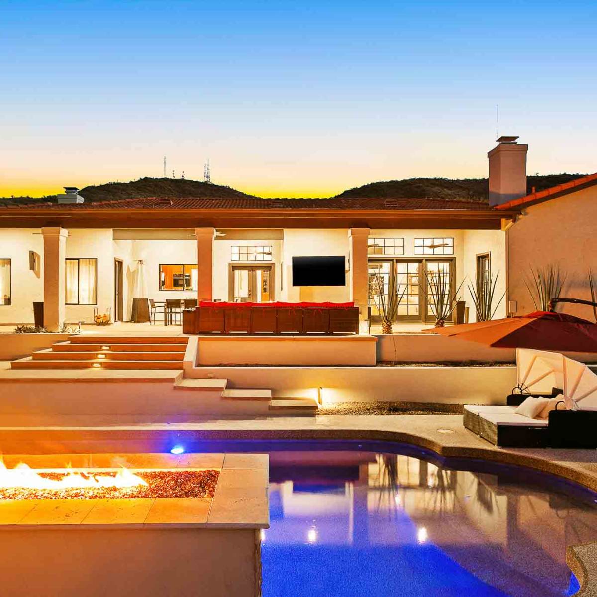 A modern single-story house with a spacious outdoor area, featuring a swimming pool, fire pit, and patio seating, under a colorful sunset sky.