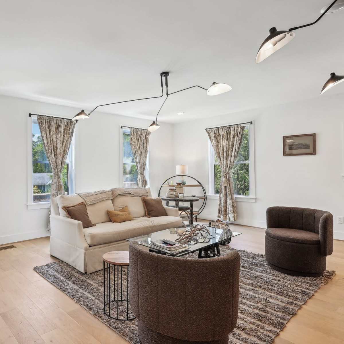 A modern living room with a white sofa, two armchairs, large windows with curtains, and a glass-top coffee table.