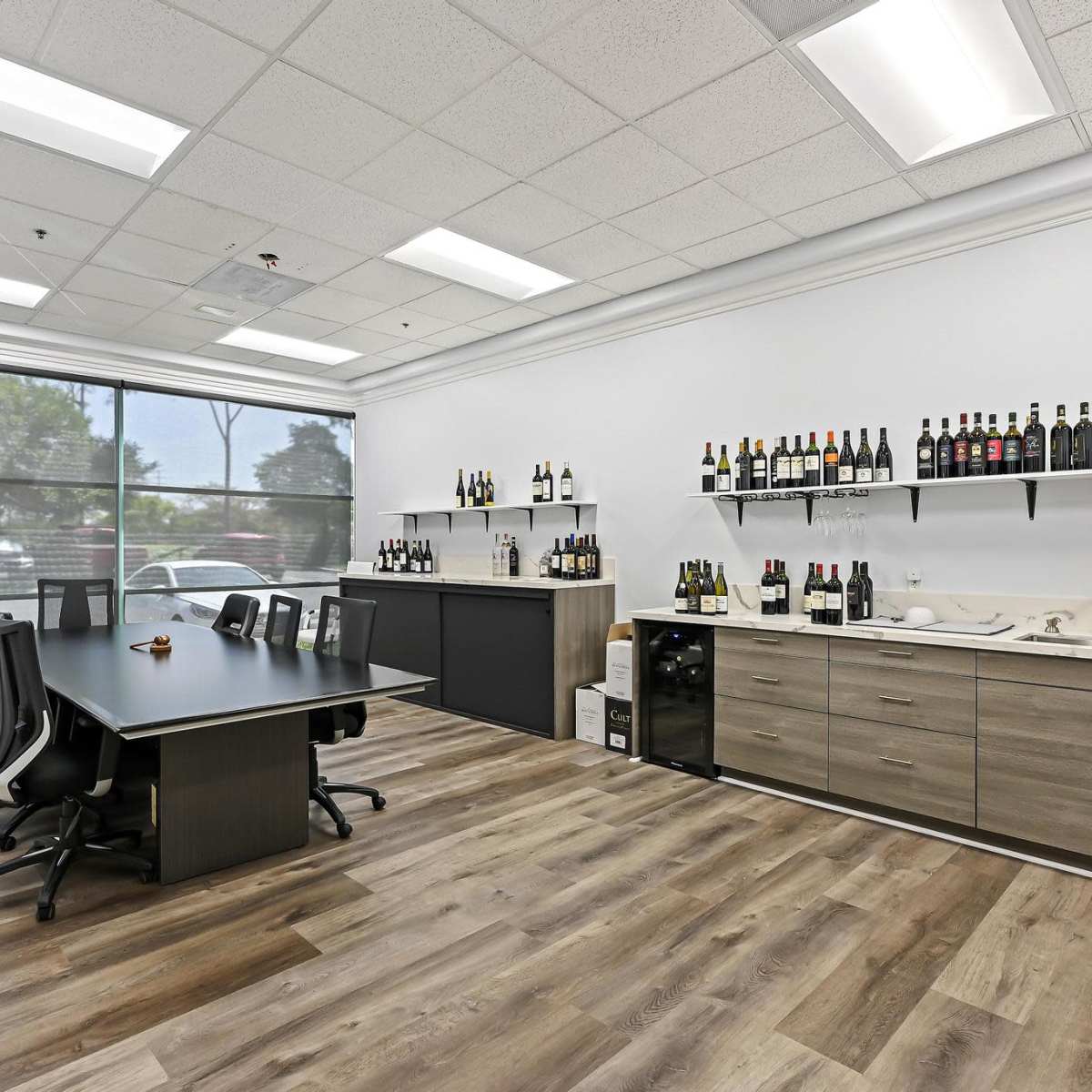 A modern office space with a large table, black chairs, a counter displaying various bottles, and a large window providing natural light.