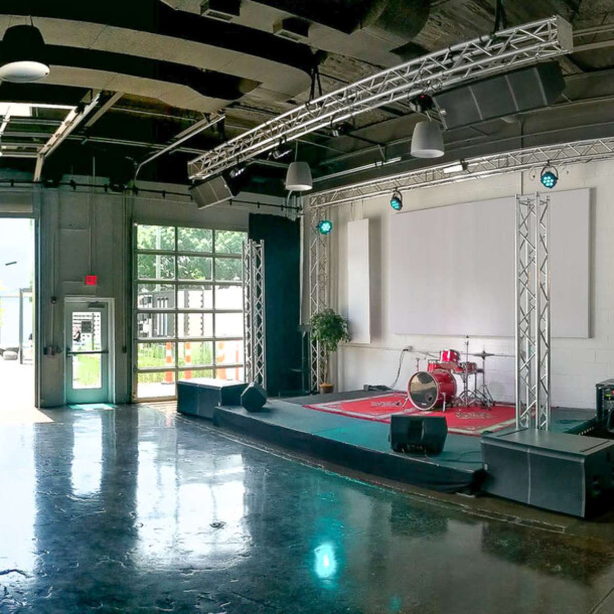 A spacious venue with a stage, drum set, and seating areas, featuring large windows that open to an outdoor view.