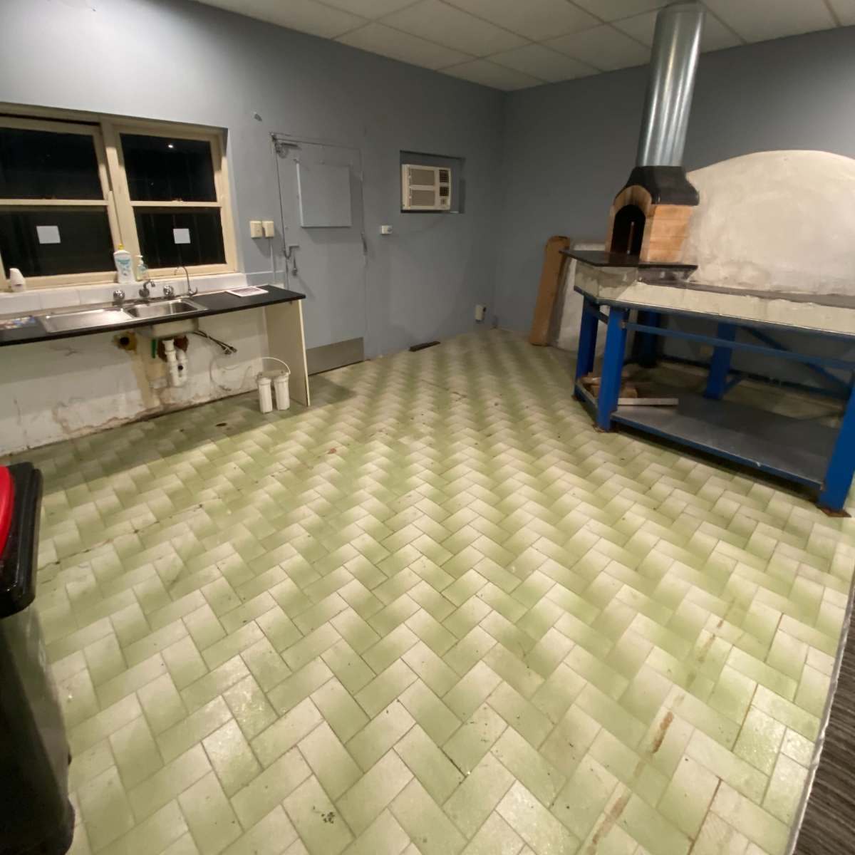 A spacious kitchen with green herringbone-patterned tiles, a wooden pizza oven, and a countertop area.