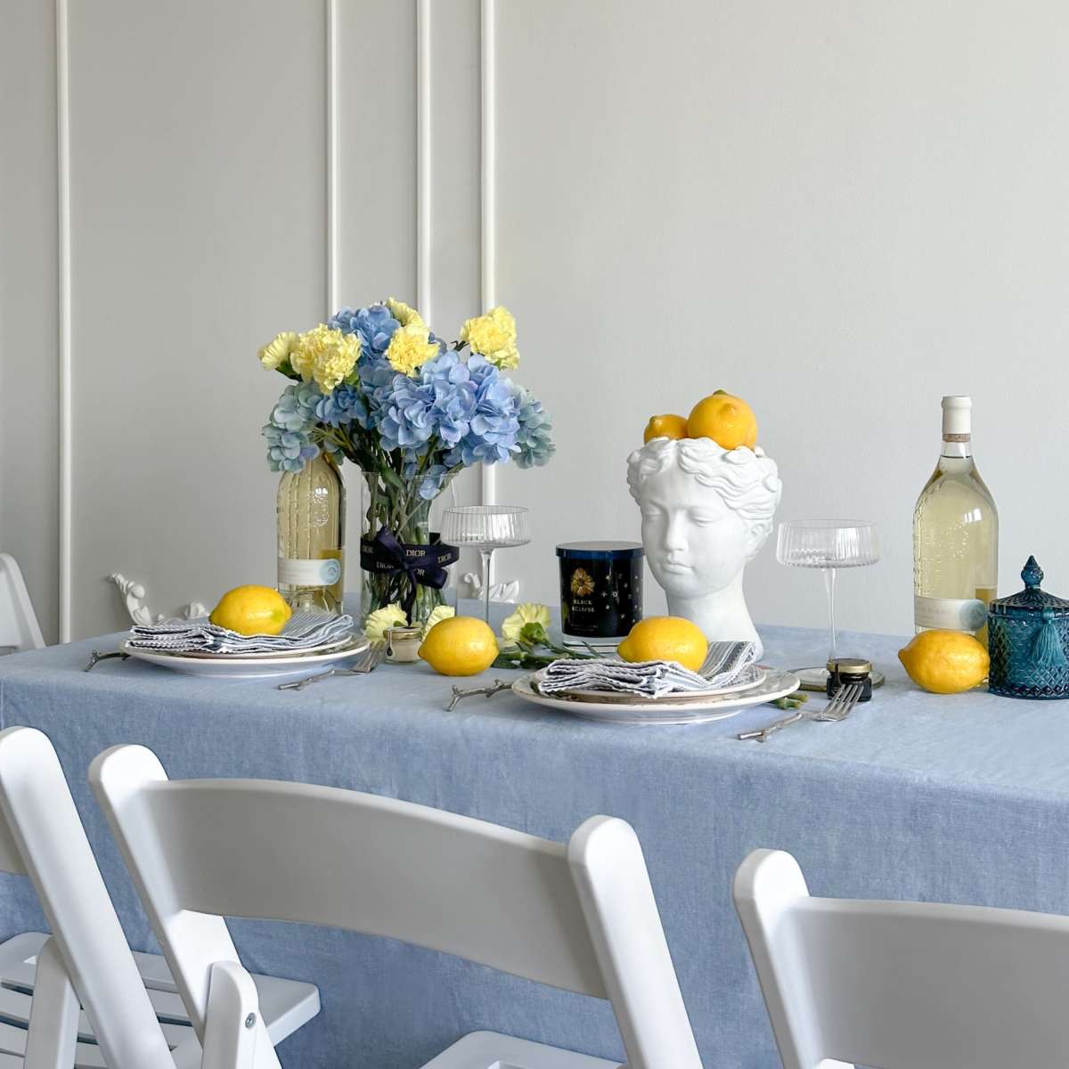 A dining table is set with a blue tablecloth, decorative flowers, and a white bust centerpiece adorned with lemons, flanked by wine bottles and glasses.