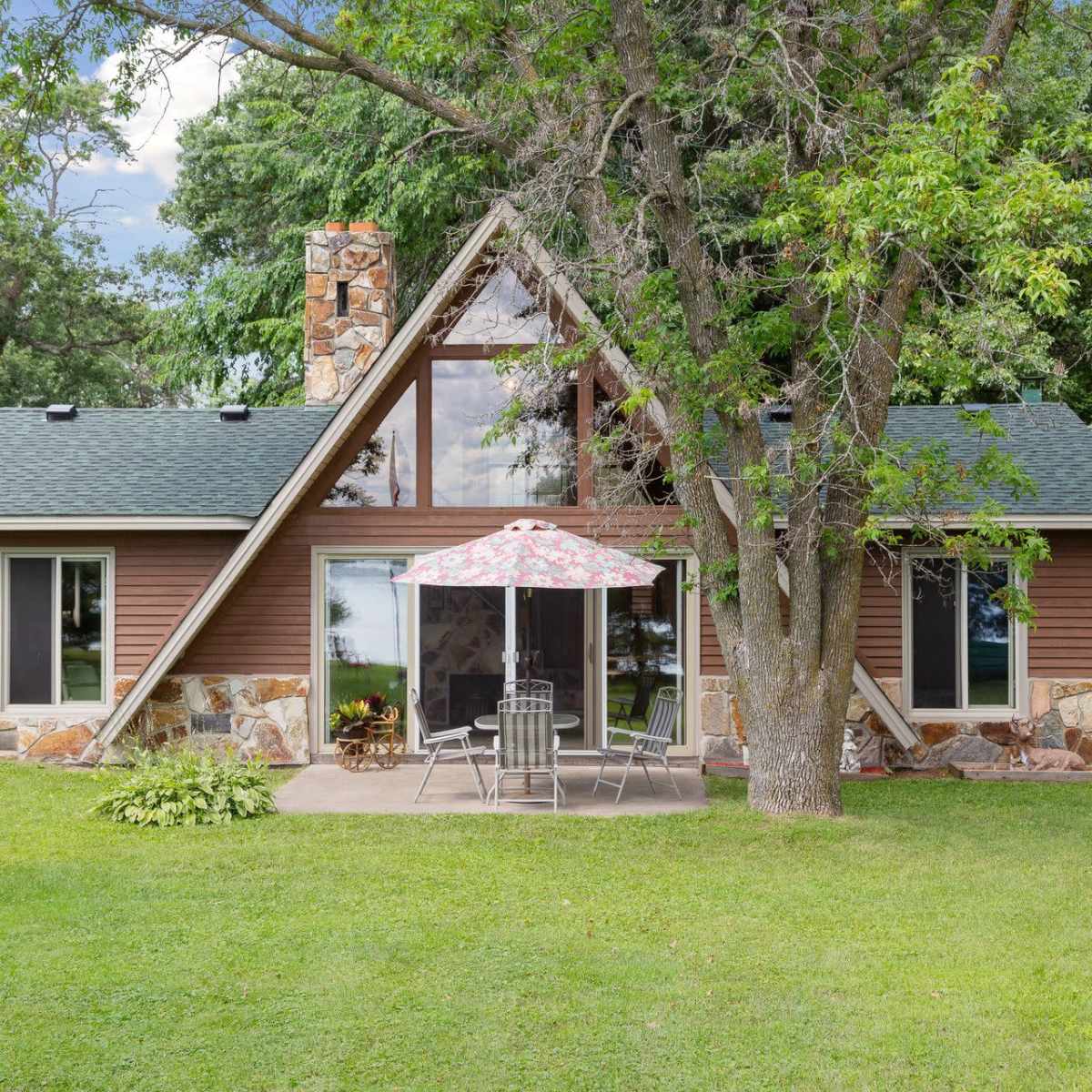 A chalet-style house with a stone chimney features a patio set and an umbrella in a grassy yard surrounded by trees.