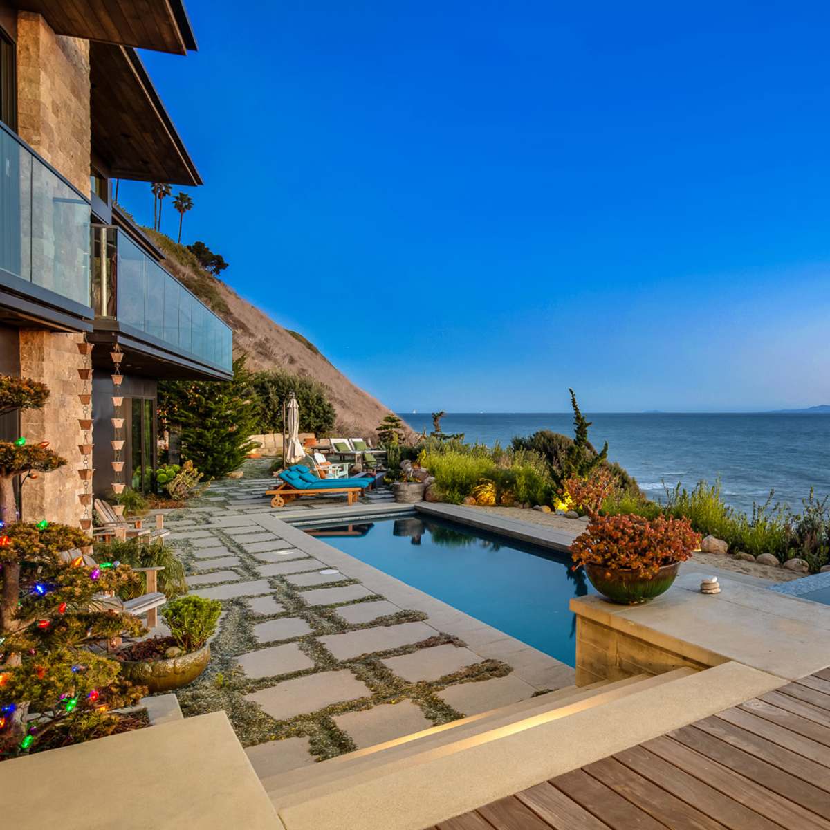A modern waterfront property with a swimming pool, hot tub, and landscaped patio overlooking the water at sunset.