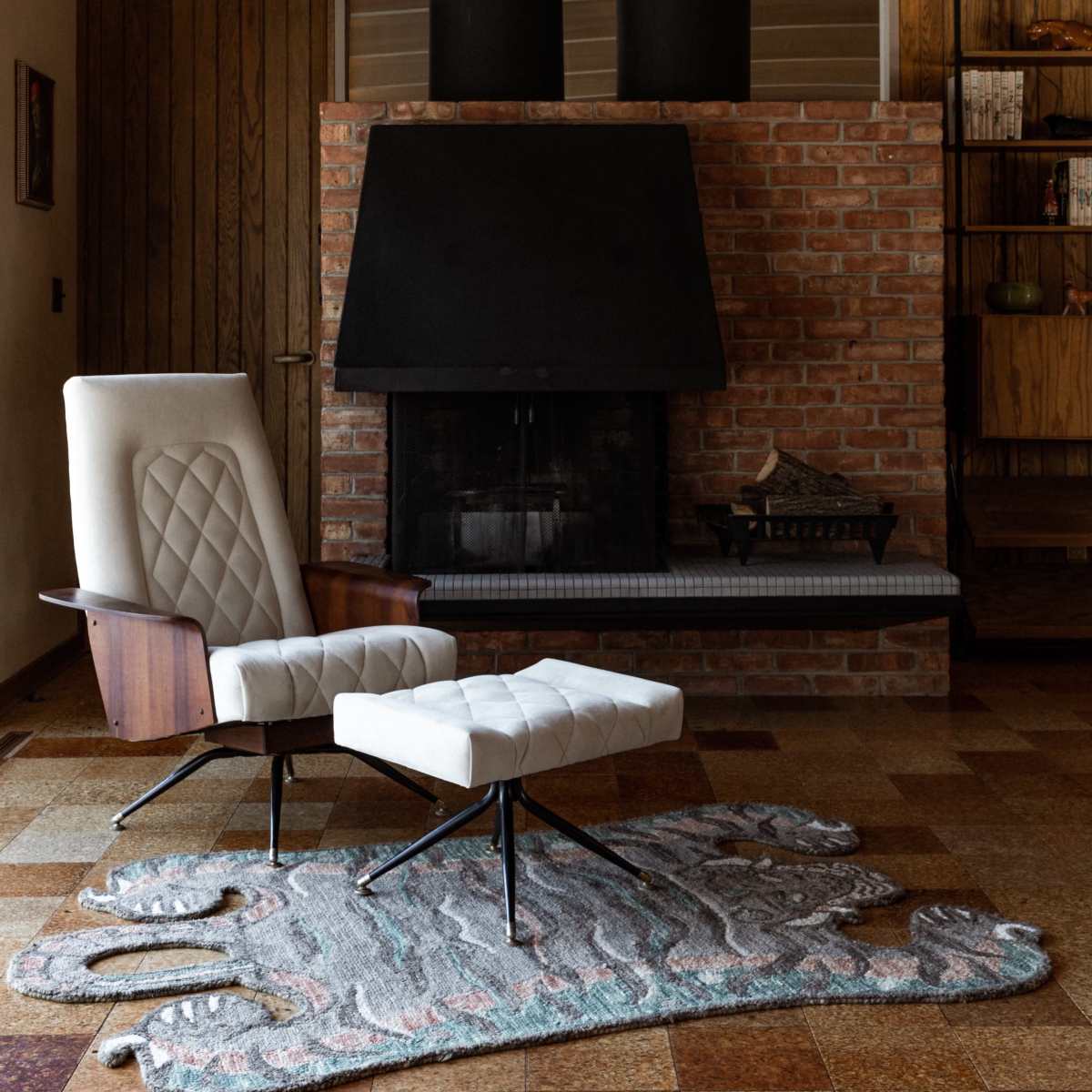 A modern lounge area featuring a white upholstered chair and ottoman in front of a brick fireplace, with wooden beams and a patterned rug on the floor.