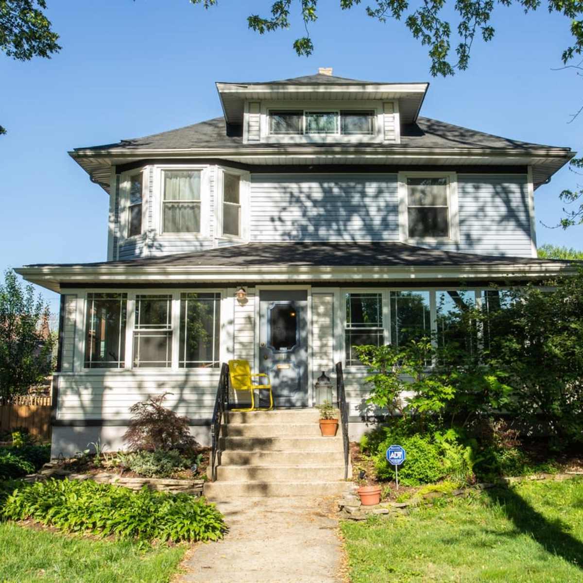 A three-story house with a front porch and steps is surrounded by a lawn and trees.