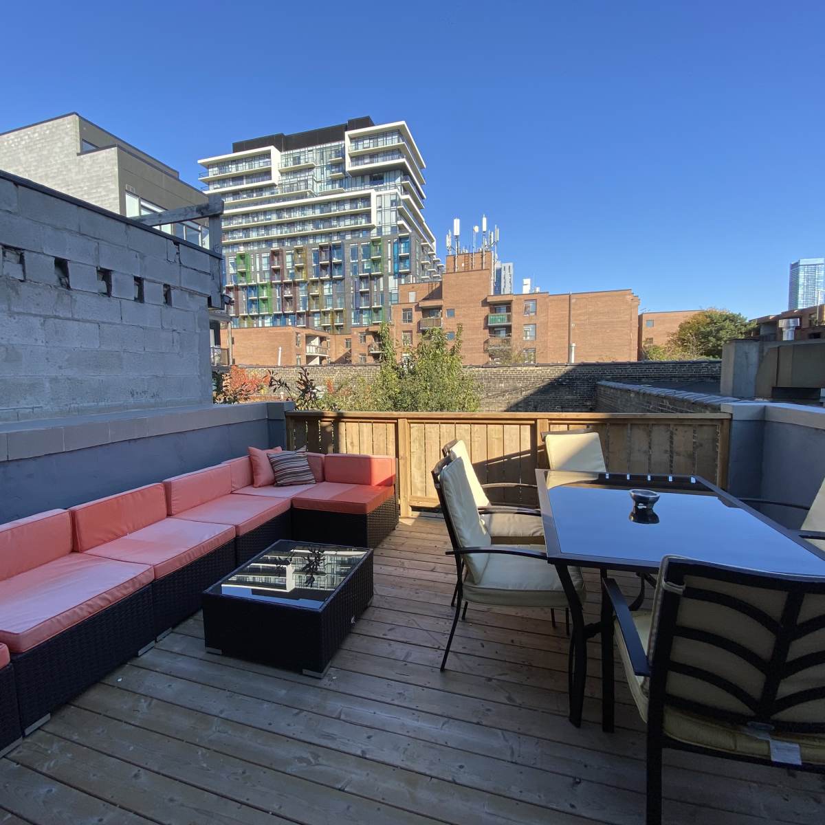 A balcony with a wooden deck featuring a seating area with orange cushions, a glass-topped table, and a view of buildings in the background.