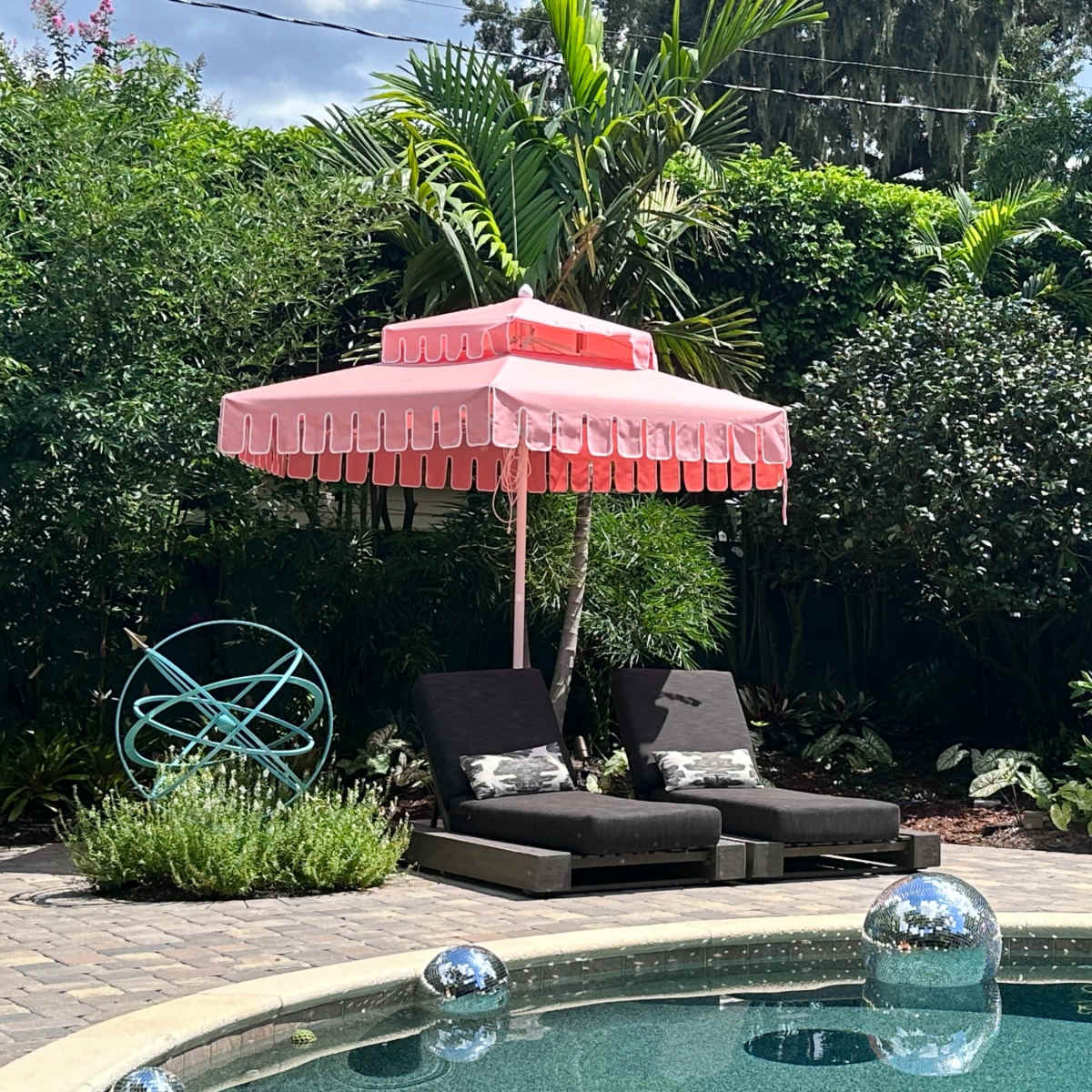 A pink umbrella shades two lounge chairs beside a shimmering pool, surrounded by lush greenery and decorative plants.