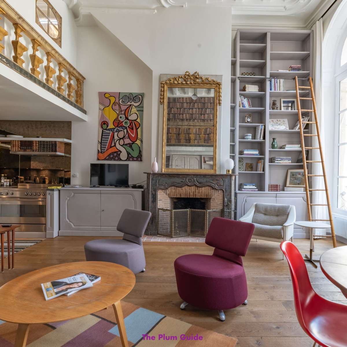 A cozy, well-decorated living space featuring a wooden floor, a fireplace, a circular dining table, colorful chairs, and large windows with natural light.