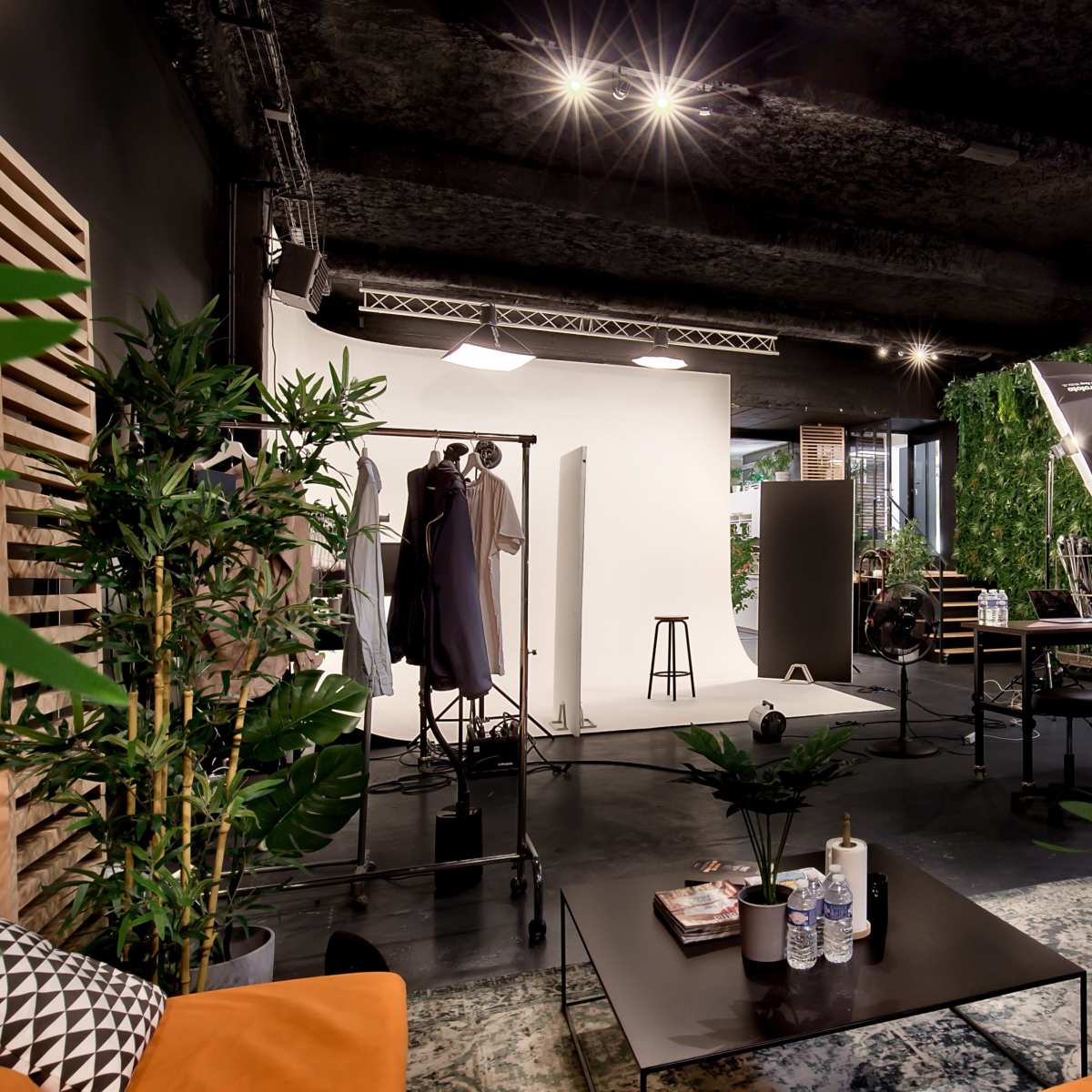 A photography studio featuring a white backdrop, various lighting equipment, and green plants alongside furniture like a table and chairs.