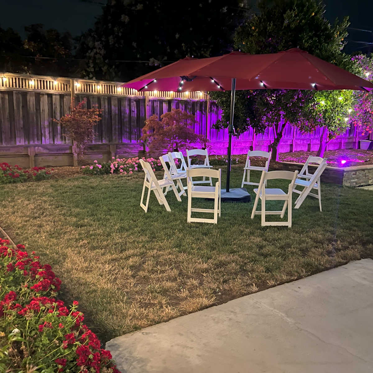 A circular arrangement of white chairs is set up under a red umbrella in a garden illuminated by purple lights.