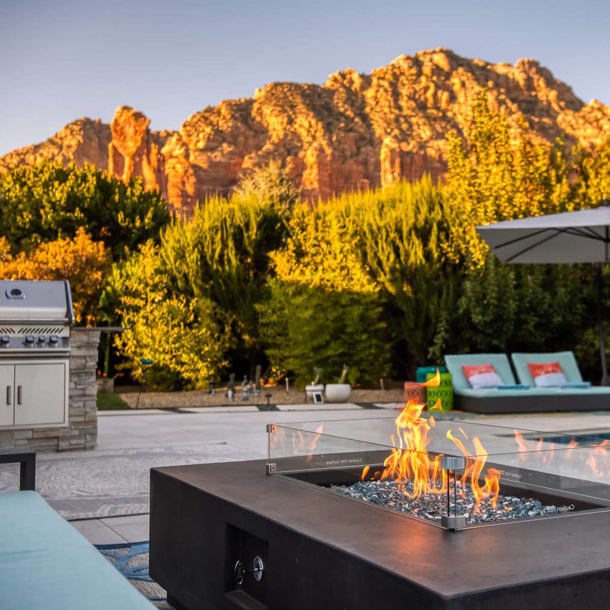 A fire pit with flames is situated in the foreground, surrounded by a stone patio, while a grill and lounge area are visible, with a backdrop of mountainous terrain and greenery.