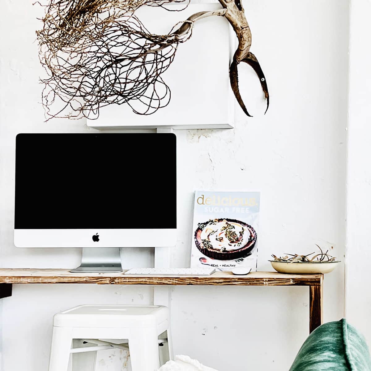 A minimalist workspace features an iMac on a wooden table with a cooking magazine and a decorative bowl, alongside a green couch.
