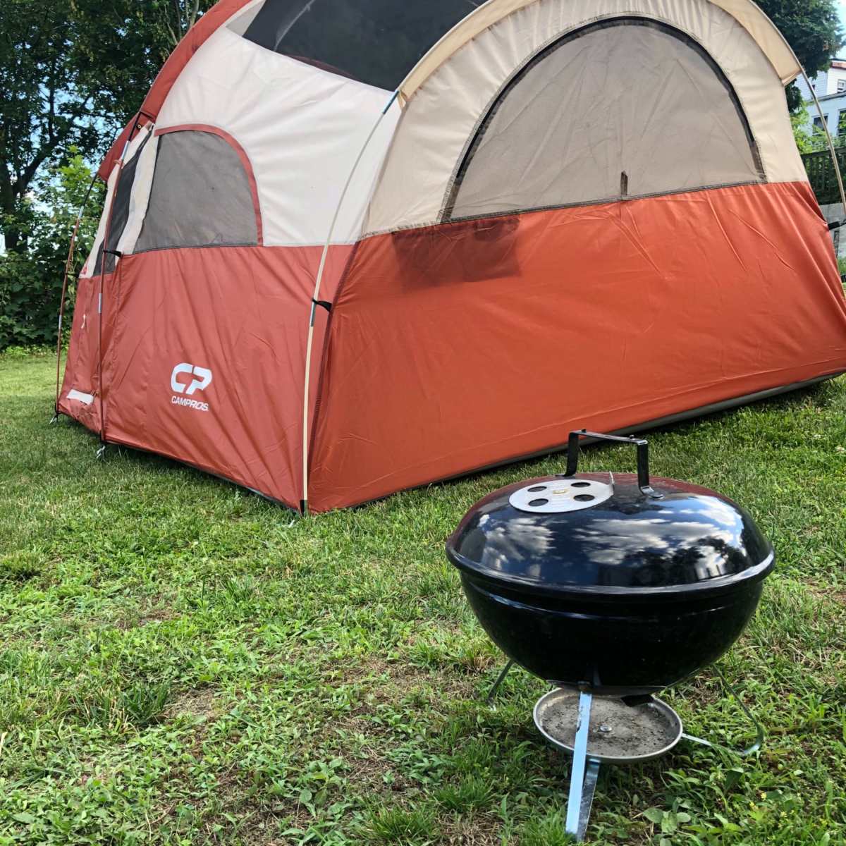 A large orange and cream tent stands on a grassy area next to a small black charcoal barbecue grill.