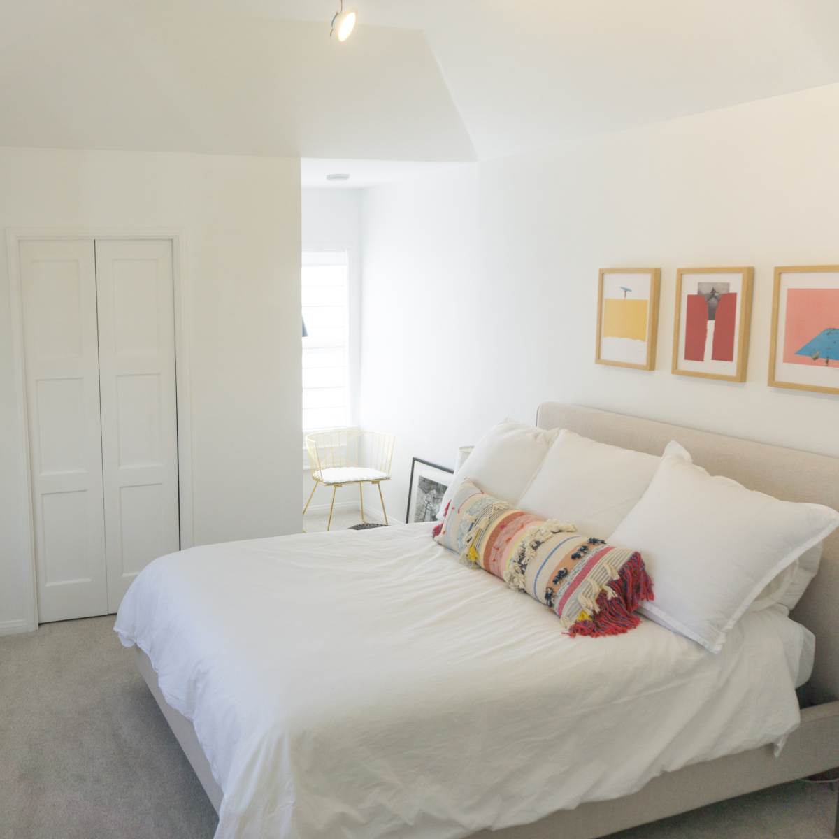 A bright bedroom features a bed with white bedding, a decorative pillow, and a small chair near a window, alongside closed double closets and framed artwork on the wall.
