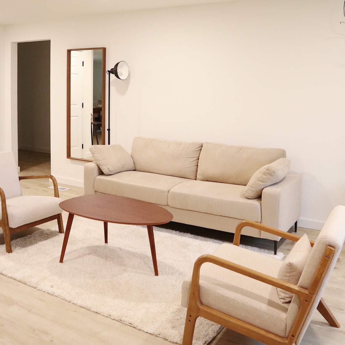 A living room features a beige sofa, two wooden armchairs, a round wooden coffee table, and a light rug.