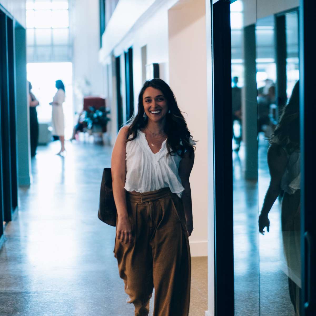 A woman in a white top and brown pants walks through a corridor with glass doors on one side.