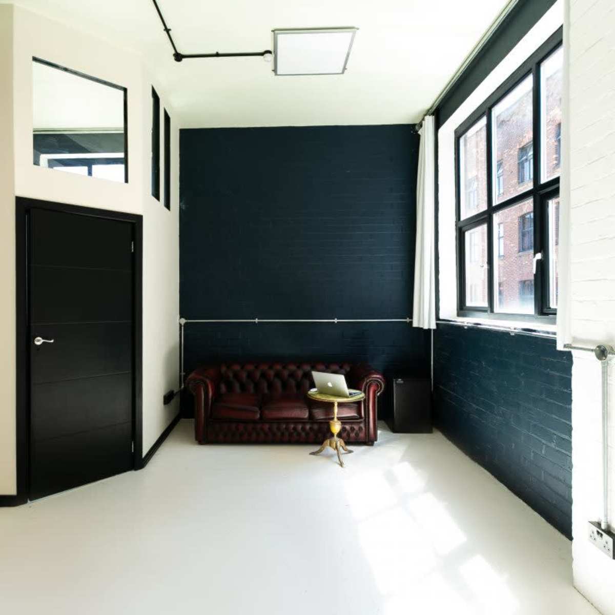 A spacious interior room with a dark wall, a brown chesterfield sofa, a small side table, and large windows allowing natural light to enter.