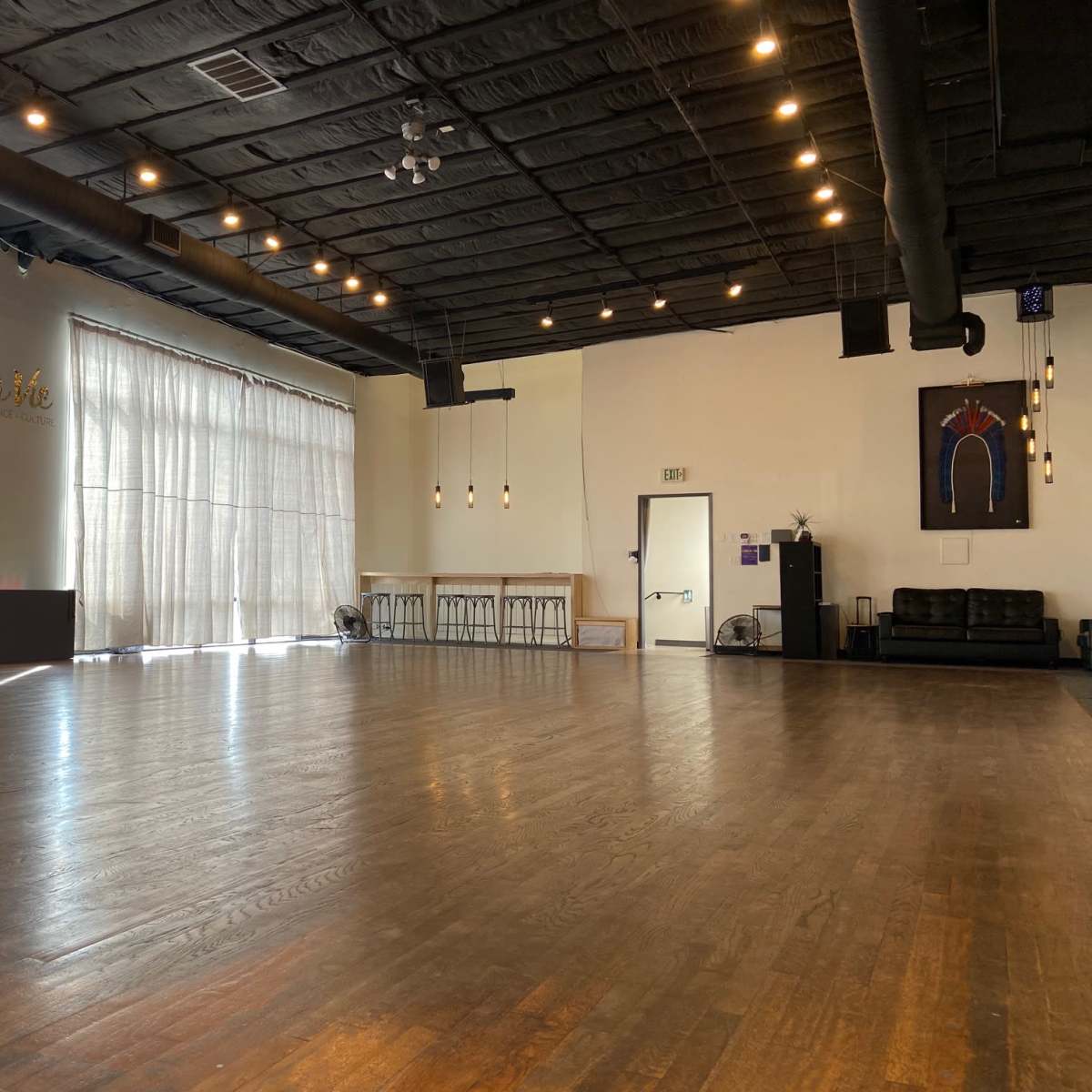An empty dance studio with large windows partially covered by sheer curtains, wooden flooring, and minimal furnishings.