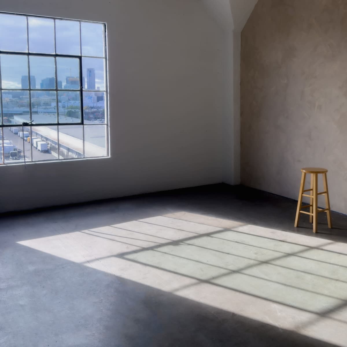 A wooden stool stands alone in a sunlit industrial room with a large window, casting a shadow on the floor.