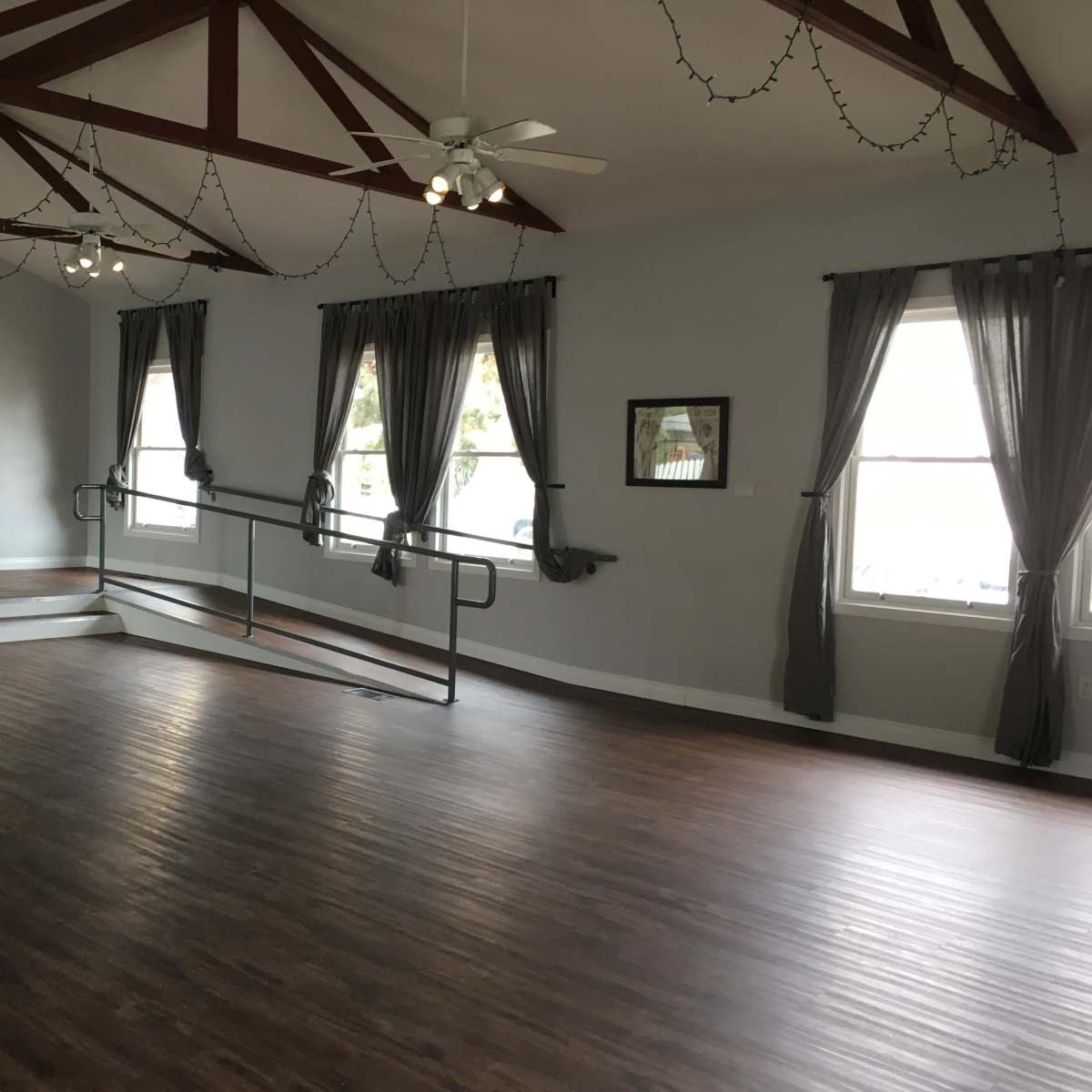 A spacious, empty room with wooden flooring, high ceilings supported by exposed beams, and large windows dressed with dark curtains.