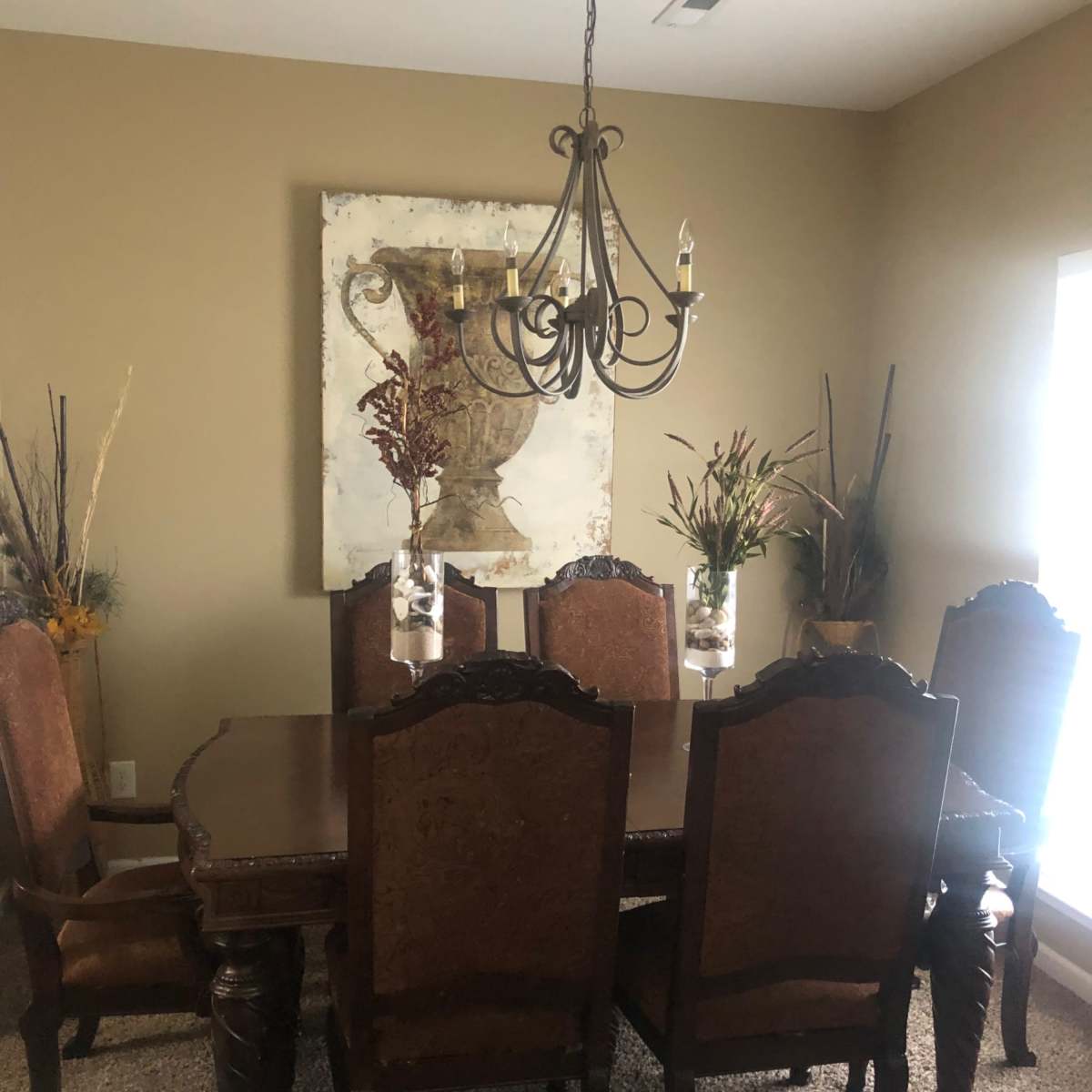 A dining room features a large wooden table surrounded by upholstered chairs, with a chandelier above and a decorative painting on the wall.