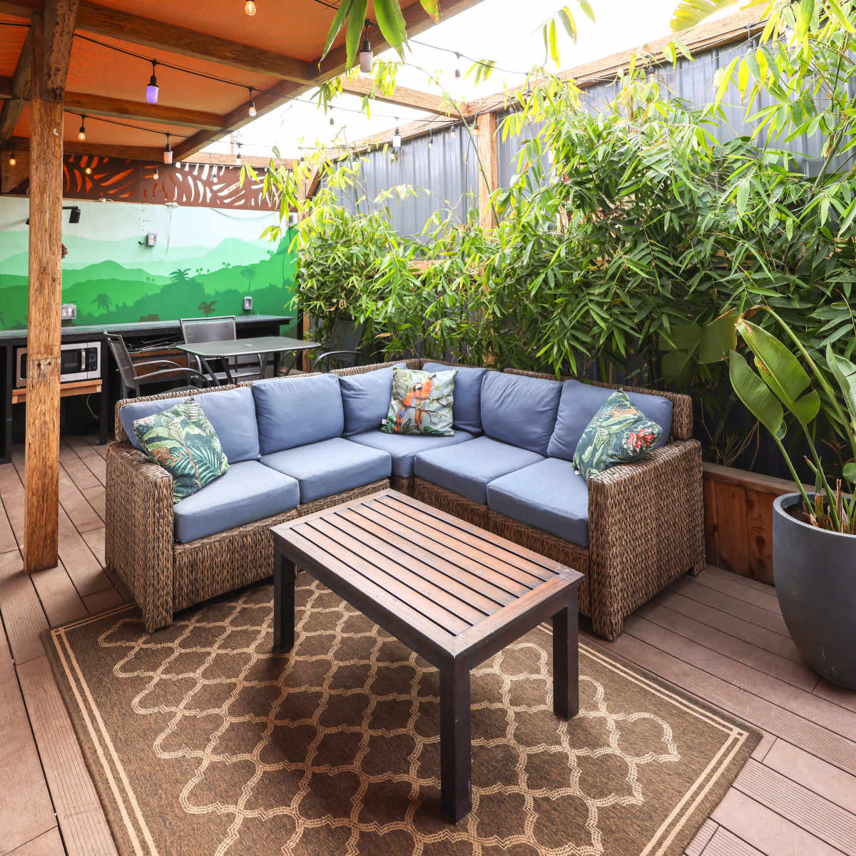 A cozy outdoor seating area features a sectional sofa with blue cushions, a wooden coffee table, and lush greenery surrounded by an orange canopy.