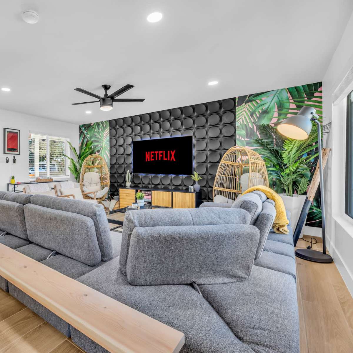 A modern living room features a large sectional sofa, a wall-mounted TV displaying the Netflix logo, and tropical-themed decor.