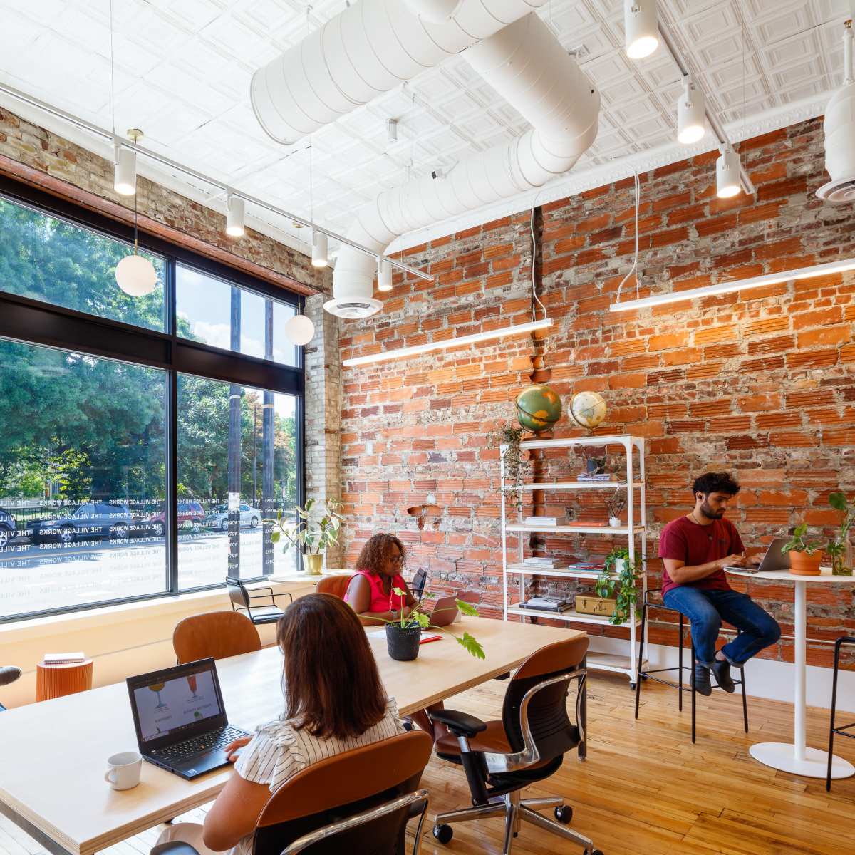 A modern office space features a mix of individuals working at a long table, chairs, and standing desks, surrounded by large windows and exposed brick walls.
