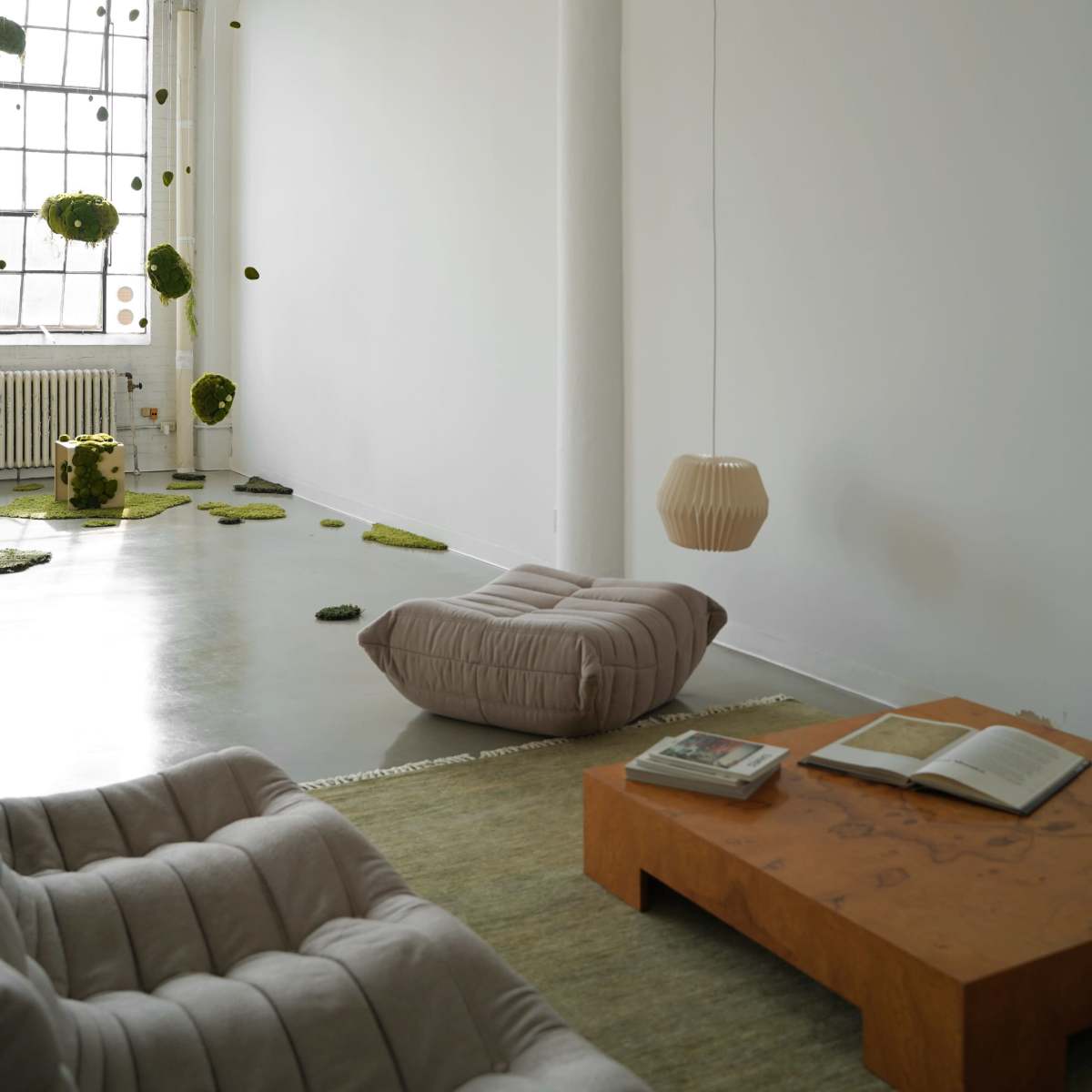 A minimalistic interior with a white wall, a cozy seating area, a wood table with an open book, and suspended decorative elements.