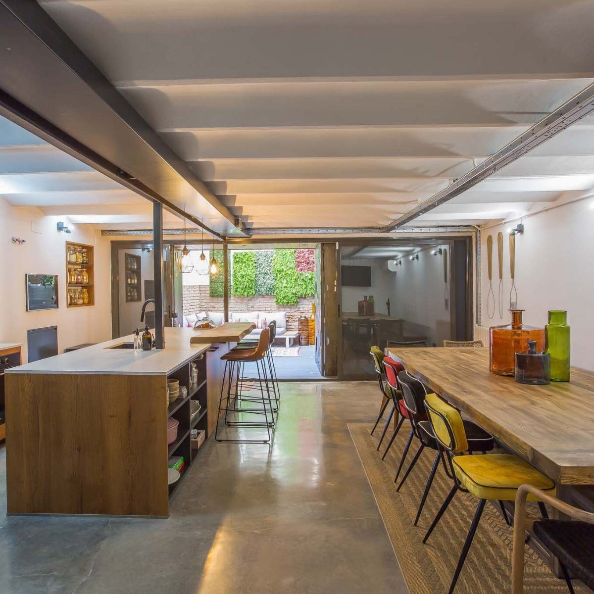 A modern kitchen and dining area with wooden furnishings, concrete flooring, and large glass doors opening to an outdoor space.