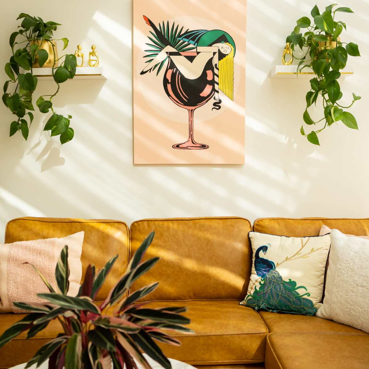 A living room with a brown leather couch, a vibrant artwork featuring a bird and cocktail, and green plants on shelves.