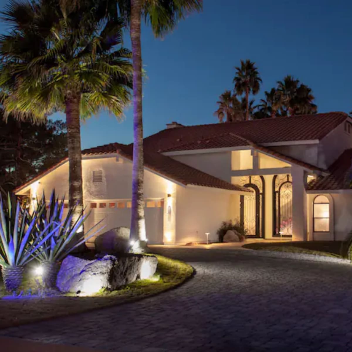 A large, well-lit house with a red-tiled roof is situated amidst palm trees and landscaped garden features under a night sky.