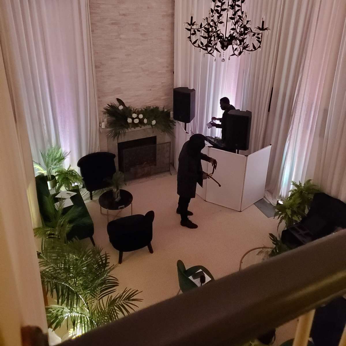 A person plays a violin in a spacious room with a fireplace, large curtains, and green plants, while another individual operates equipment at a DJ booth.