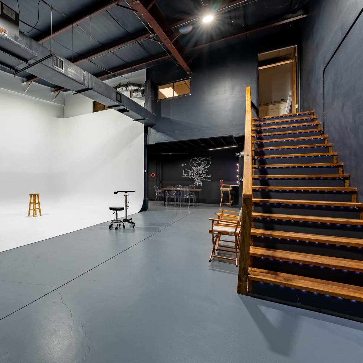 A spacious studio with a white backdrop, wooden stairs leading to an upper level, and various furniture arranged for shooting or meetings.