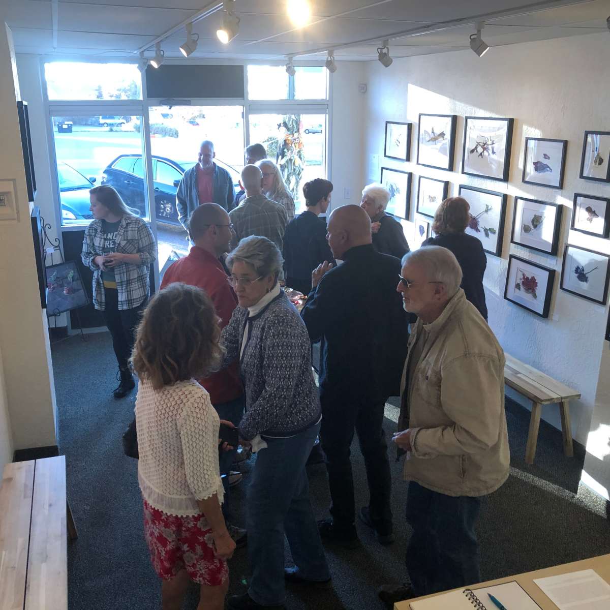 A group of people stands inside an art gallery, surrounded by framed artworks on the walls, with some conversing and others observing the pieces.