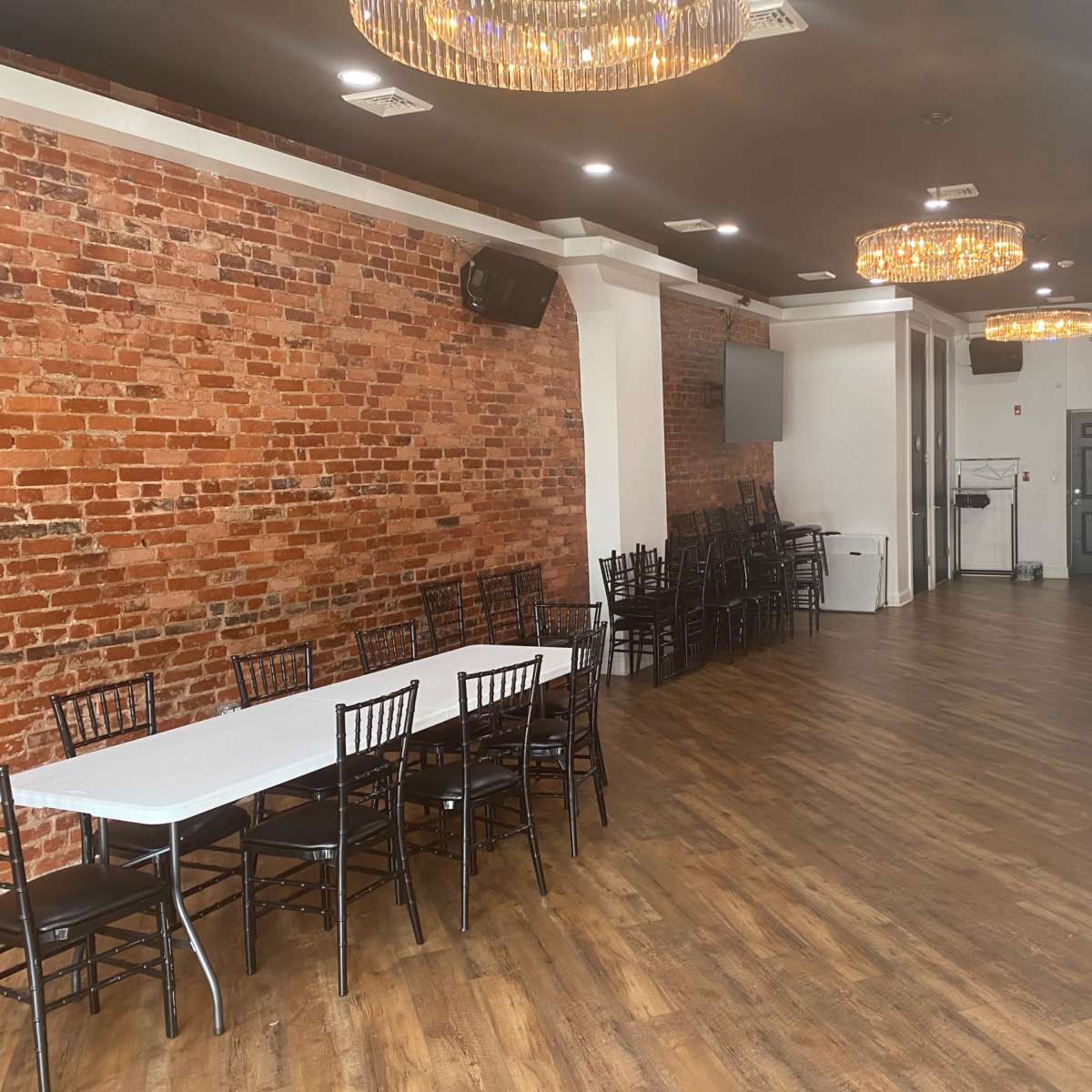A spacious room with exposed brick walls, a long white table with black chairs, and a modern kitchen area at the far end.