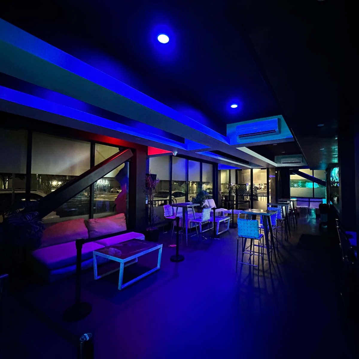 A dimly lit lounge with blue and red lighting, featuring tables, chairs, and large windows overlooking an urban view at night.