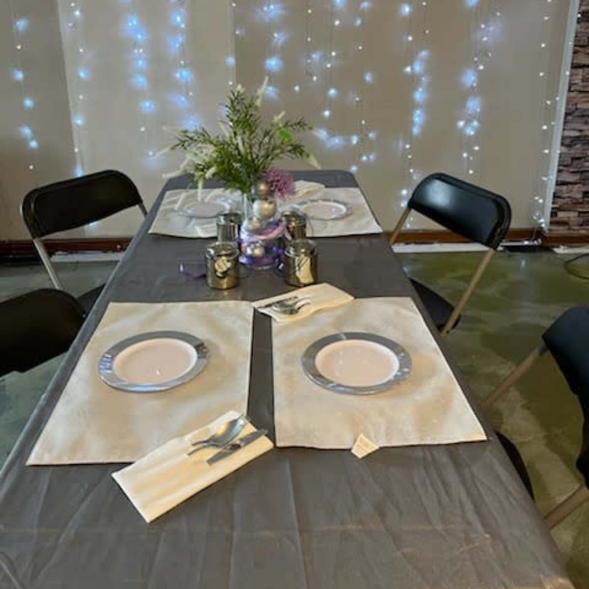A dining table is set with four plates and decorative elements, surrounded by chairs and illuminated by strands of twinkling lights on the wall.