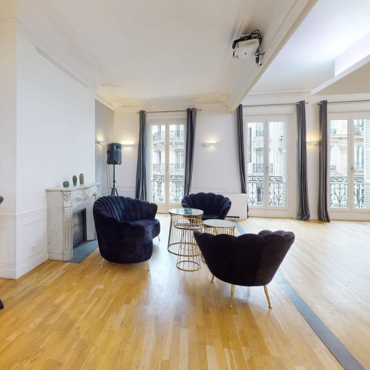 An elegant room with parquet flooring, featuring two black armchairs, a round table, a tall cocktail table, and large windows overlooking an exterior balcony.
