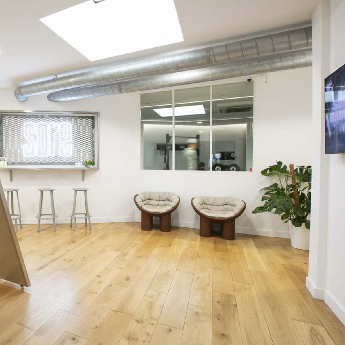 A modern office space features wooden flooring, a metal table with stools, and two plush chairs next to a large window displaying the word "SODE."