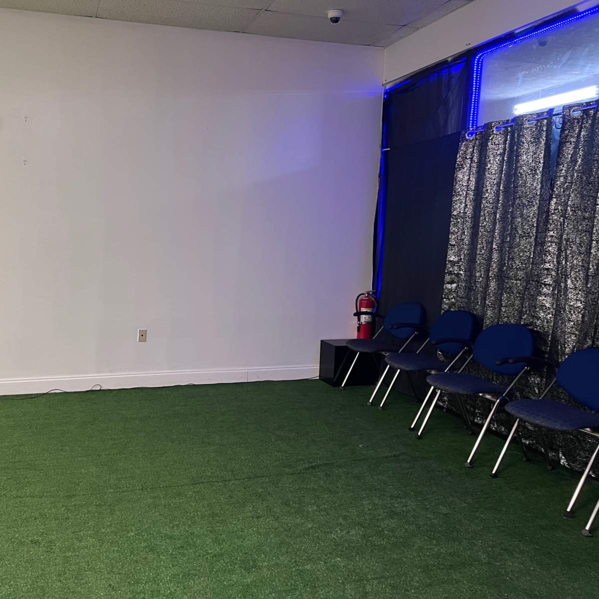 An empty room with green artificial turf, a row of blue chairs, and a fire extinguisher against the wall.