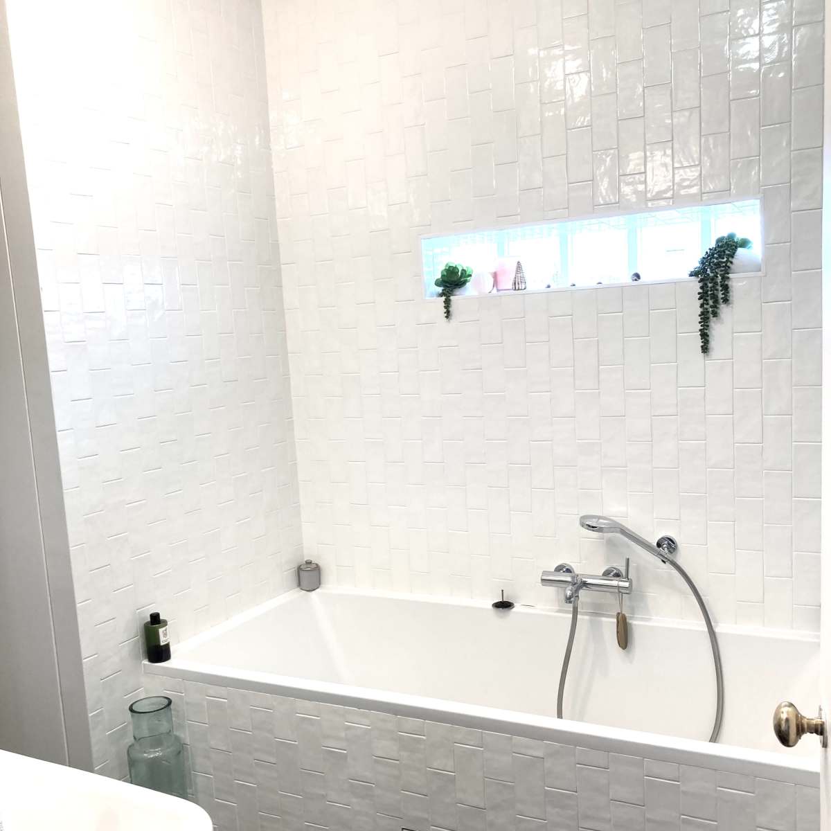 A modern bathroom featuring a white tiled wall and a bathtub with a shower fixture, complemented by a small window with decorative plants.