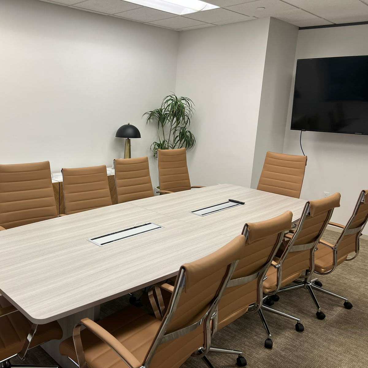 A modern conference room with a long table surrounded by eight brown chairs, against a backdrop of a sideboard and a TV on the wall.