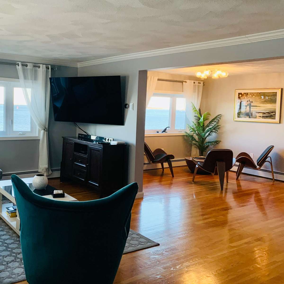 The living room features a large window overlooking the ocean, with a gray couch and a blue accent chair arranged around a coffee table and entertainment center.